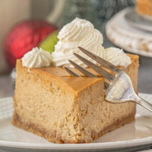 “Close-up of a slice of gingerbread cheesecake with whipped cream swirls on top, resting on a white plate with a fork ready to take a bite.”