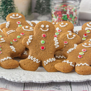 Christmas gingerbread man cookies decorated with white icing and red and green buttons on a white platter with holiday sprinkles in the background.