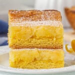 Two stacked lemon chess bars on a white plate, topped with powdered sugar, showing a golden cake mix crust and gooey lemon-cream cheese filling, with a lemon twist garnish on the side.