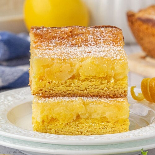 Two stacked lemon chess bars on a white plate, dusted with powdered sugar, showing a golden crust and gooey lemon-cream cheese filling.