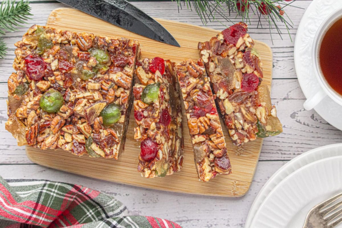 Sliced old-fashioned fruitcake on a wooden cutting board, packed with pecans, red and green candied cherries, and dates, next to a cup of tea and holiday plaid napkin.