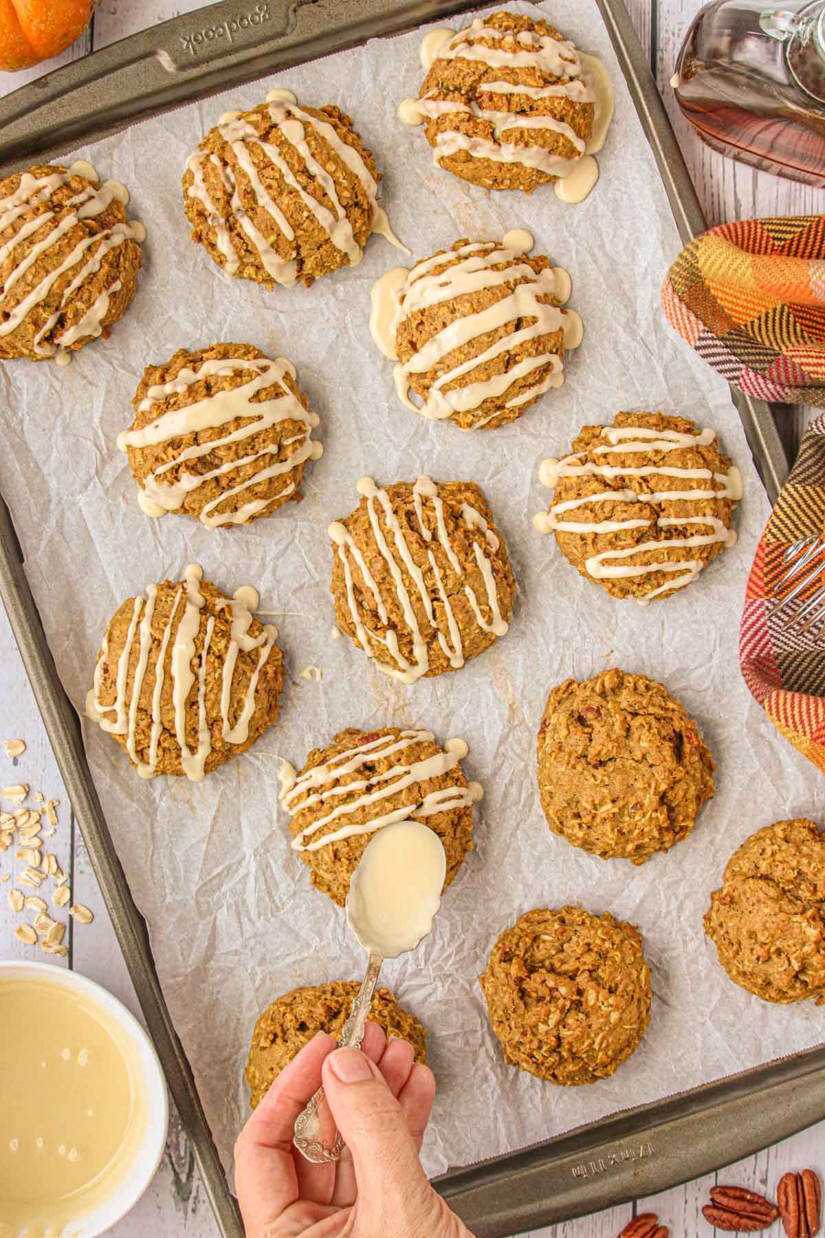 Freshly baked pumpkin oatmeal cookies on a parchment-lined baking sheet, being drizzled with icing from a spoon.