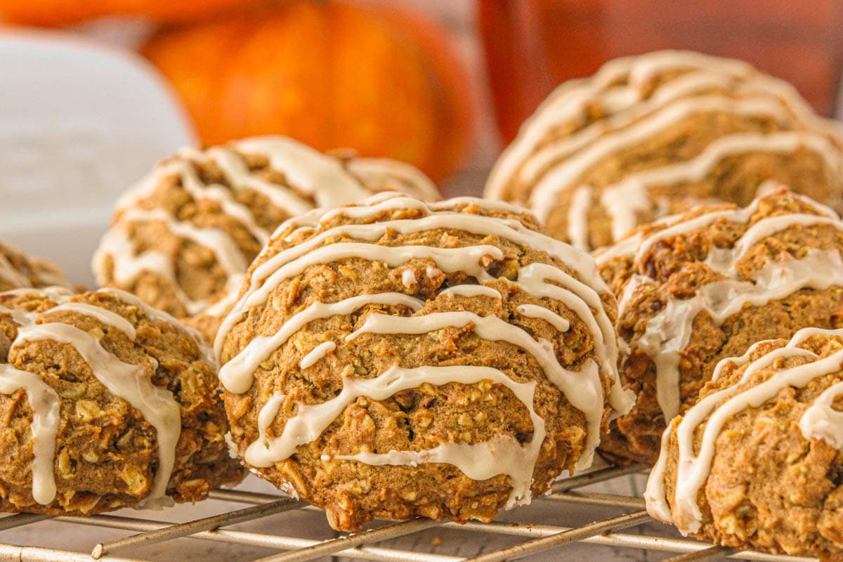 Pumpkin oatmeal cookies with icing drizzle cooling on a wire rack, with small pumpkins in the background for a fall touch.