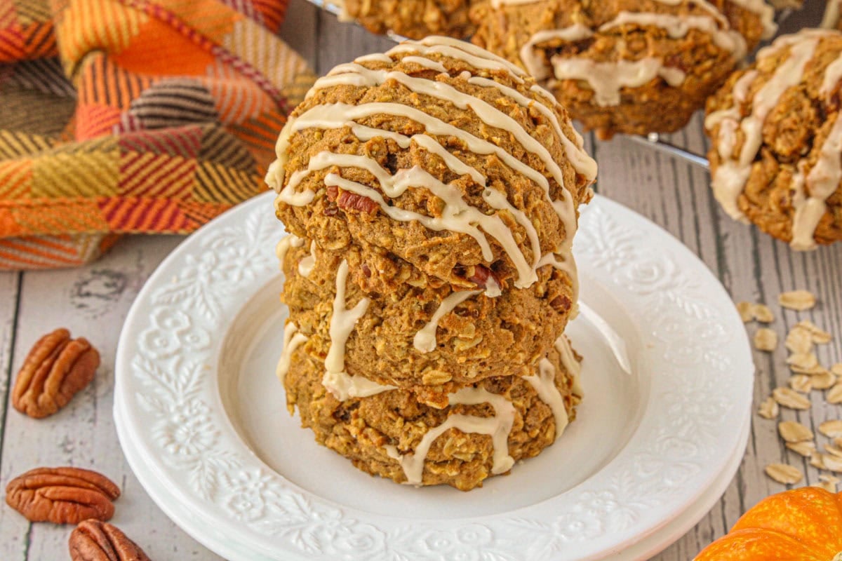 A stack of chewy pumpkin oatmeal cookies with maple icing on a white plate surrounded by fall decor.