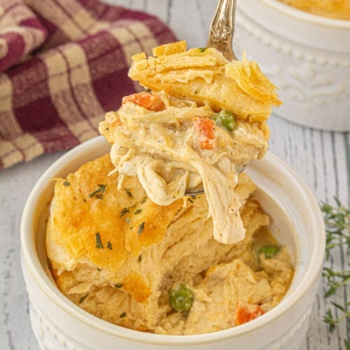 Close-up of freezer-friendly chicken and gravy casserole in a white ramekin, with a spoon lifting creamy chicken, peas, carrots, and flaky golden biscuit topping.