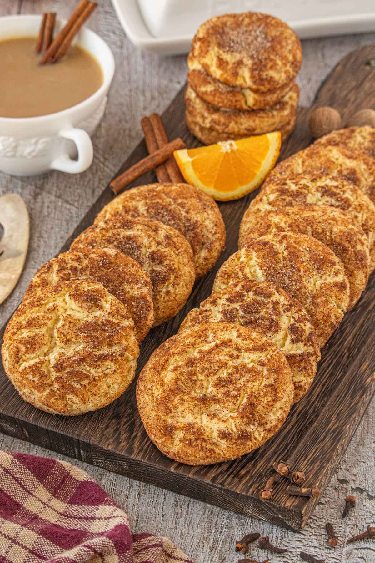 A rustic wooden board filled with chai snickerdoodle cookies coated in cinnamon sugar, arranged in neat rows next to a few stacked cookies, cinnamon sticks, whole cloves, and a wedge of orange. A cup of chai sits in the background, creating a warm and cozy baking scene.