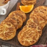 Rows of chai snickerdoodle cookies arranged on a dark wooden board, garnished with cinnamon sticks and an orange wedge, with a cup of chai tea behind them.