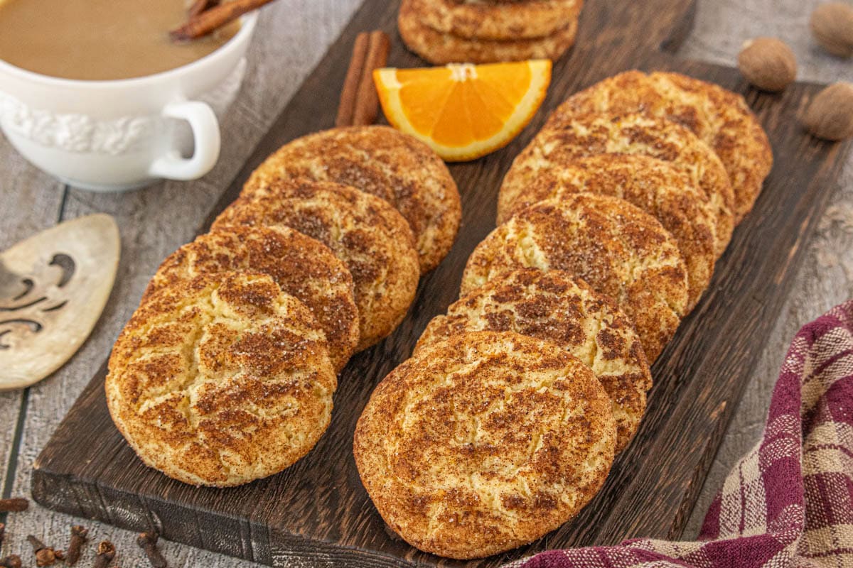 Rows of chai snickerdoodle cookies arranged on a dark wooden board, garnished with cinnamon sticks and an orange wedge, with a cup of chai tea behind them.