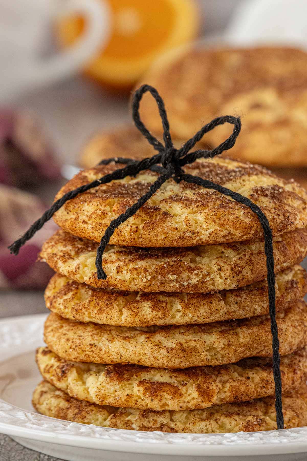A stack of chai snickerdoodle cookies tied with a black twine bow, showing the cinnamon sugar crackle on each cookie, with more cookies and an orange slice blurred in the background.