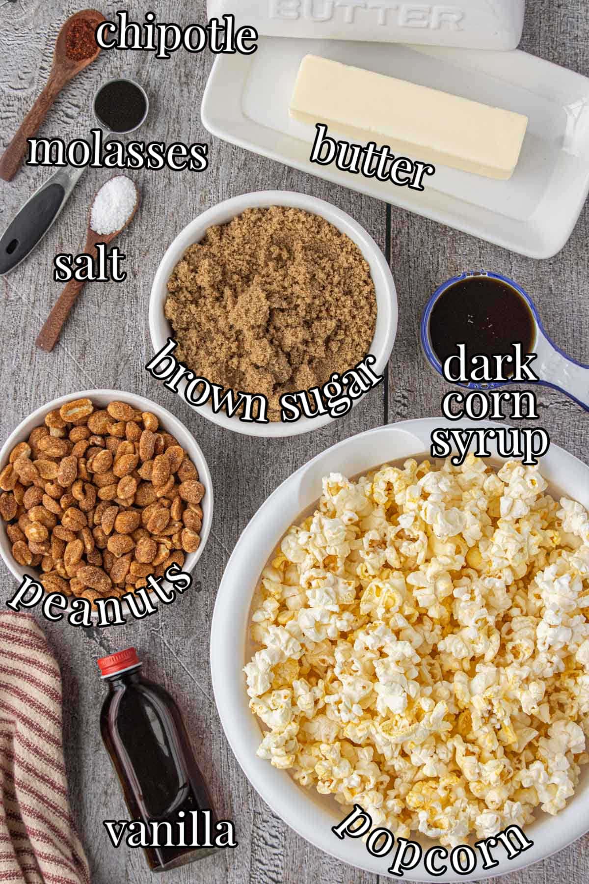 Overhead view of ingredients for homemade Cracker Jack popcorn including butter, brown sugar, molasses, dark corn syrup, chipotle powder, peanuts, vanilla, and popped popcorn in white bowls on a rustic table.