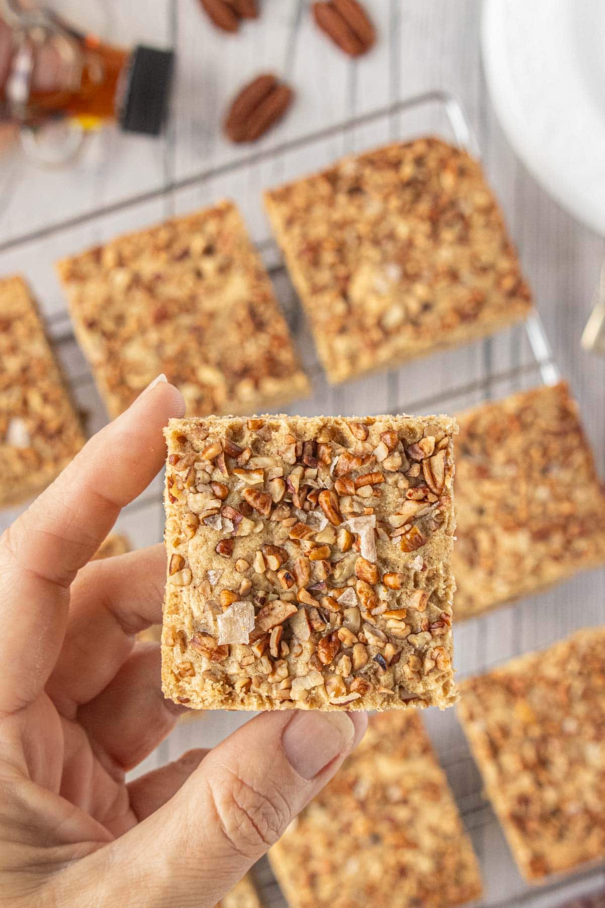 Fresh maple pecan blondie bar, golden and nutty with flaked smoked salt on top, held up over a cooling rack-soft inside, crunchy on top, pure fall comfort.
