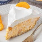 Slice of orange cream pie with mandarin orange bits and a fluffy white topping, served on a white plate with a silver fork and orange peel curls for garnish.