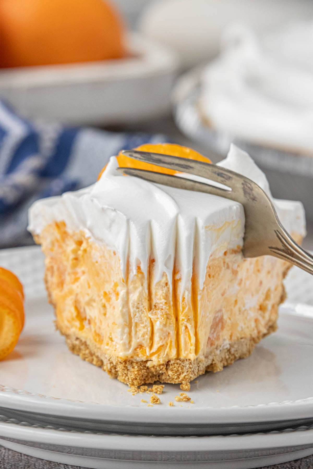 A thick, frosty slice of orange fluff pie with whipped topping and mandarin orange garnish, sitting on a white plate as a fork presses into the creamy layer.