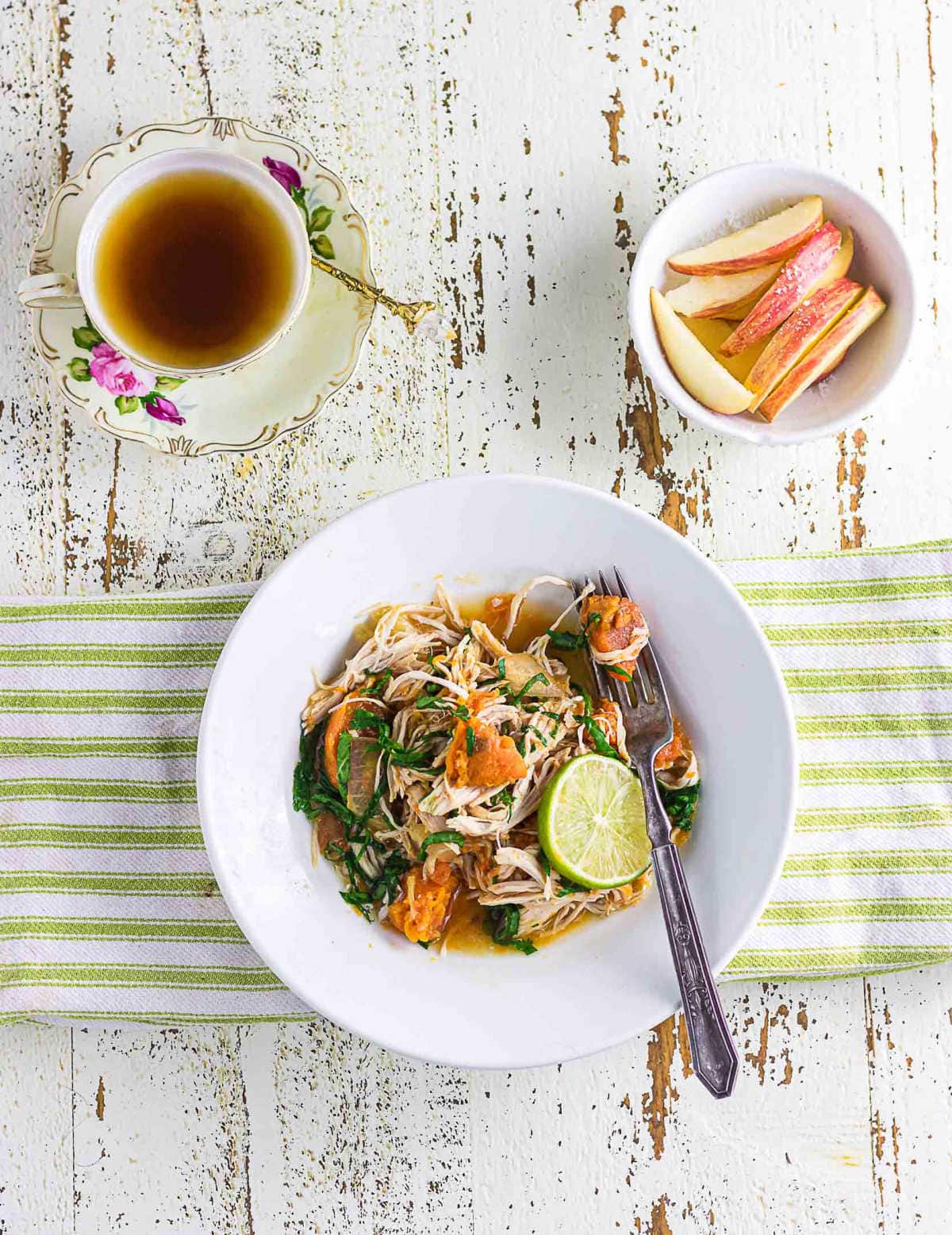 A white bowl filled with shredded chicken, sweet potatoes, and wilted spinach in a light broth, topped with a slice of lime. The bowl rests on a green-striped cloth on a distressed white table, with a vintage teacup of hot tea and a small bowl of sliced apples nearby.