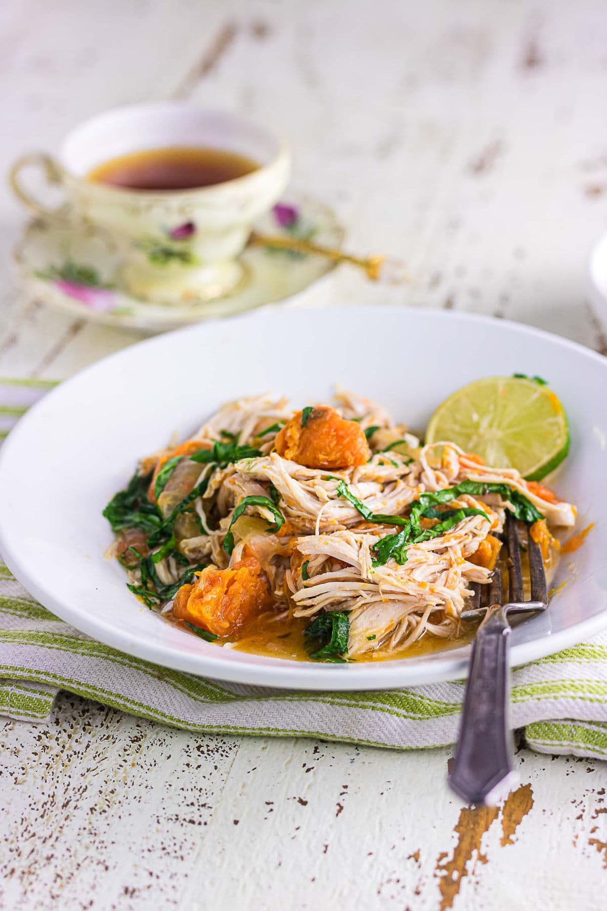A white bowl of chicken stew with sweet potatoes and spinach on a white farmhouse table.