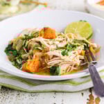 A white bowl filled with shredded chicken, tender sweet potatoes, and wilted spinach in a light broth, topped with a slice of fresh lime. The bowl sits on a green-striped cloth beside a vintage fork, with a cup of tea and sliced apples in the background on a rustic white table.