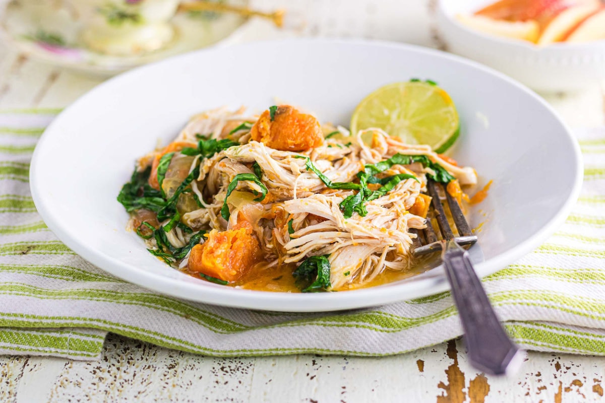 A white bowl filled with shredded chicken, tender sweet potatoes, and wilted spinach in a light broth, topped with a slice of fresh lime. The bowl sits on a green-striped cloth beside a vintage fork, with a cup of tea and sliced apples in the background on a rustic white table.