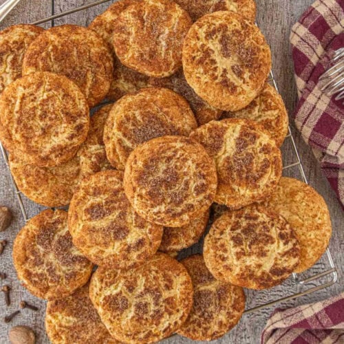 A large batch of chai snickerdoodle cookies piled onto a cooling rack, showing golden cinnamon-speckled tops and rustic cracked surfaces.
