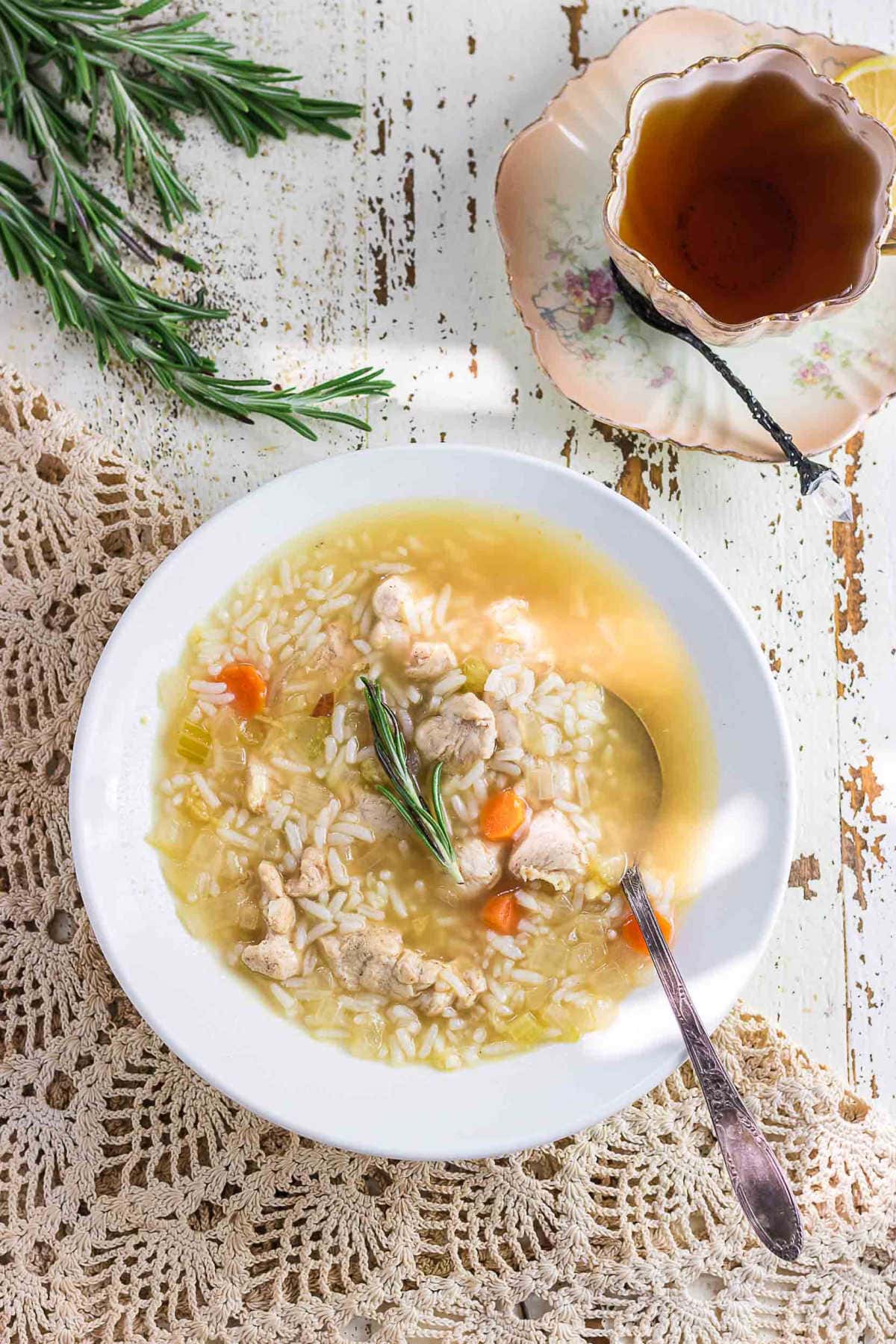 Bowl of ginger chicken and rice soup on a rustic white table with a sprig of rosemary and a vintage teacup beside it — cozy, comforting, and softly lit like a quiet afternoon in the kitchen.