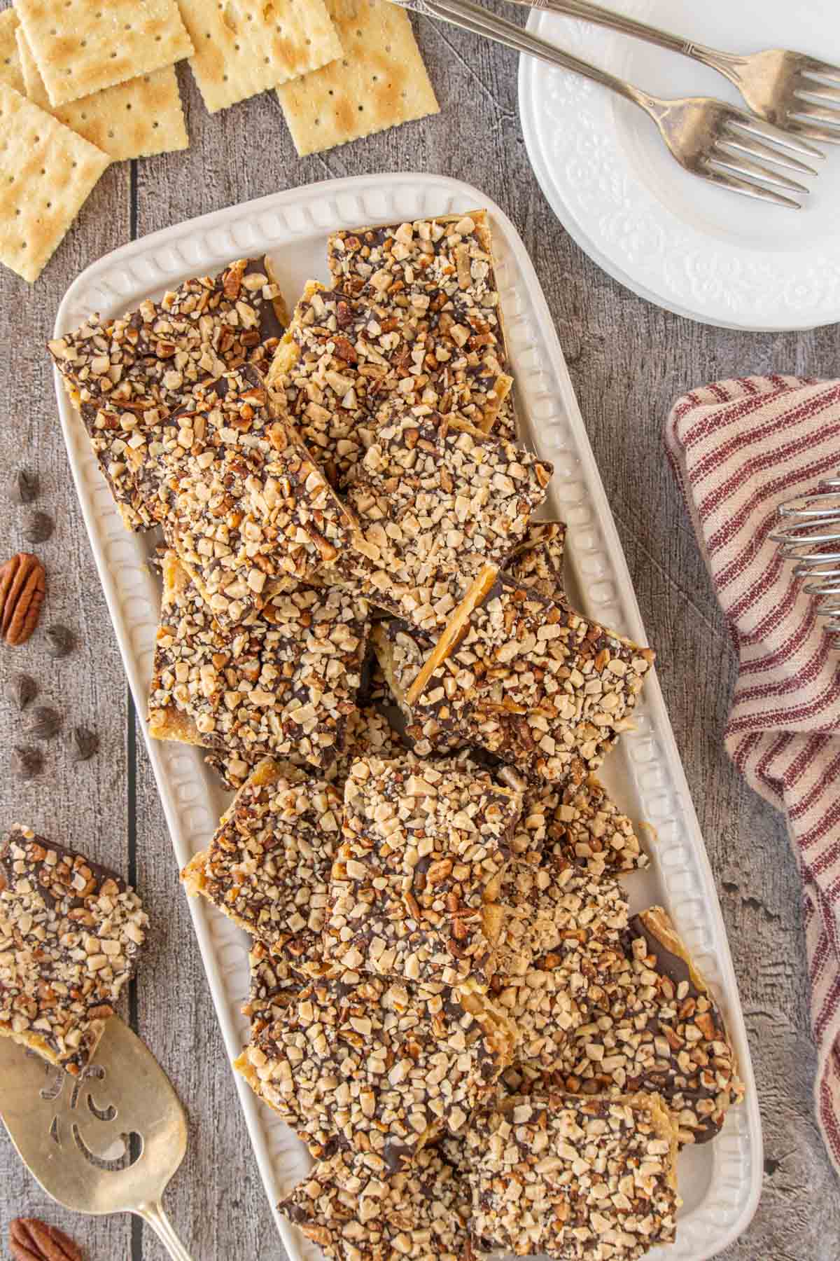 An overhead shot of a rectangular platter filled with neatly arranged saltine toffee squares, surrounded by extra crackers, forks, and a striped kitchen towel.