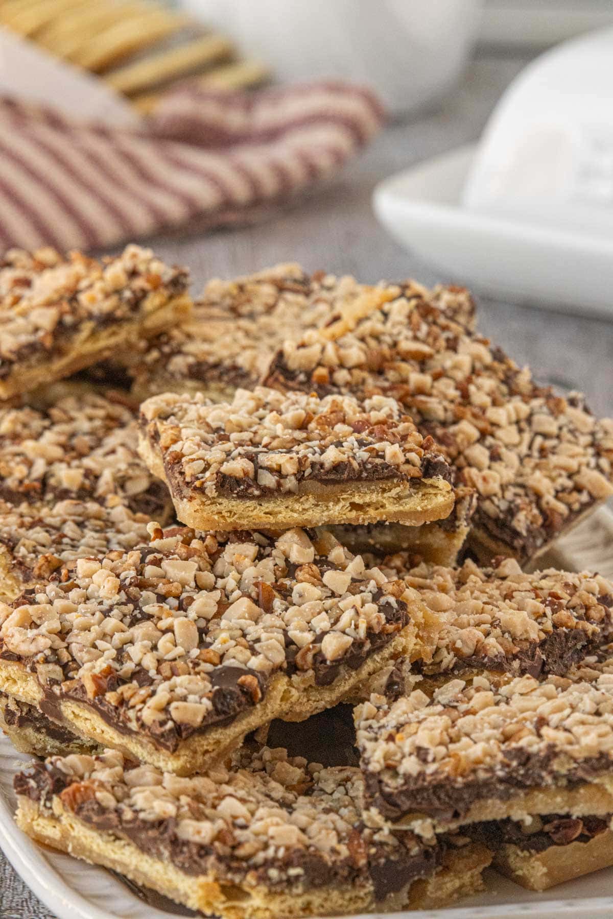 A stack of saltine toffee pieces on a white plate, highlighting the layers of cracker, caramel, chocolate, and chopped nuts with a soft, blurred background.