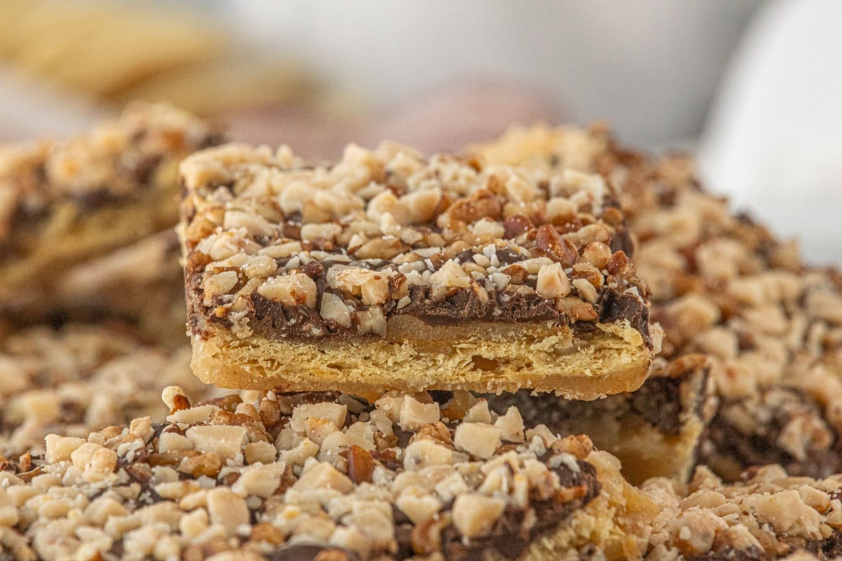 A close-up of a single piece of saltine toffee held above a platter, showing the crisp cracker base, golden caramel layer, melted chocolate, and chopped pecan topping with sharp detail.