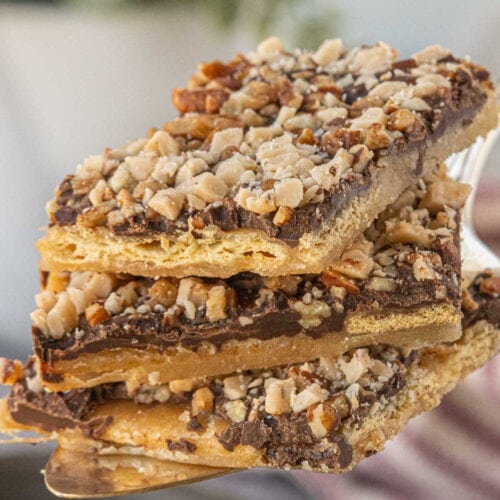 Close-up stack of saltine toffee pieces on a serving spatula, showing the cracker base, caramel layer, melted chocolate, and chopped nut topping.