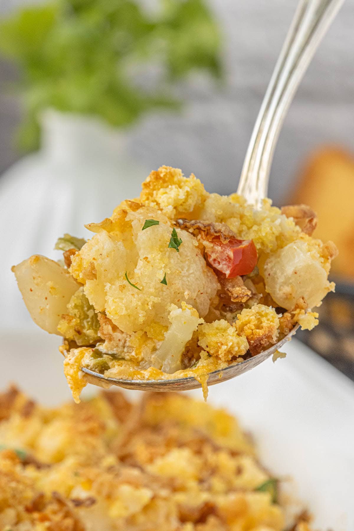 Spoonful of cheesy cauliflower casserole topped with golden cornbread crumbs, crispy onions, and colorful bell peppers in a white dish.