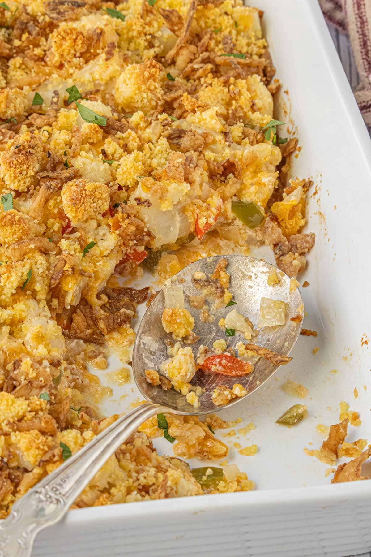 Southern cauliflower casserole with cornbread topping in a white baking dish, showing creamy texture and crispy browned edges with a vintage silver serving spoon.