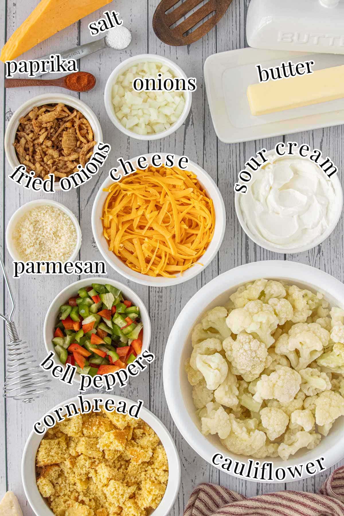 Overhead view of cauliflower casserole ingredients including cauliflower florets, cornbread crumbs, cheddar cheese, sour cream, butter, onions, and colorful bell peppers on a rustic gray surface.