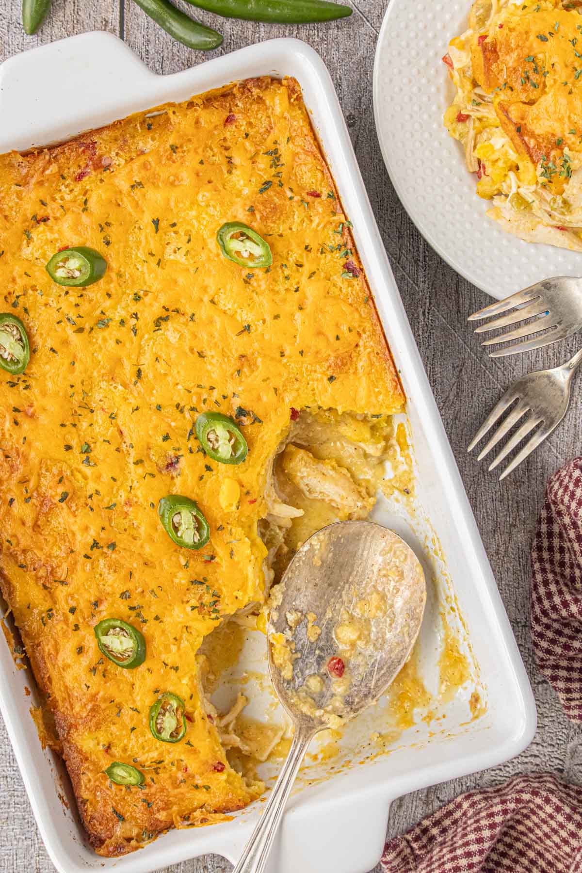 A white baking dish filled with cheesy chicken and chile cornbread casserole, topped with sliced jalapeños; one corner scooped out to show the creamy chicken and corn filling underneath.