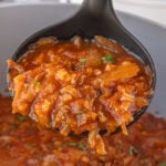 A black ladle lifting a hearty scoop of cabbage roll soup from a large skillet, showing tender cabbage, ground beef, tomatoes, and rice in a rich red broth.