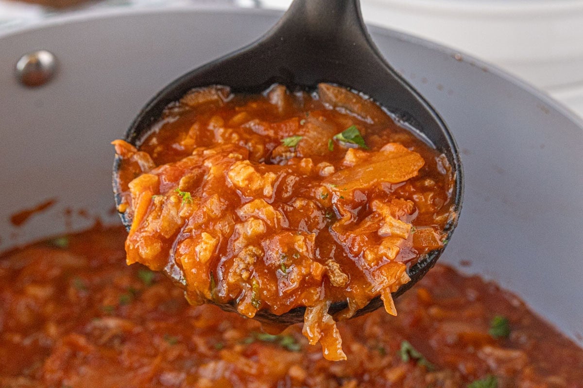 A black ladle lifting a hearty scoop of cabbage roll soup from a large skillet, showing tender cabbage, ground beef, tomatoes, and rice in a rich red broth.