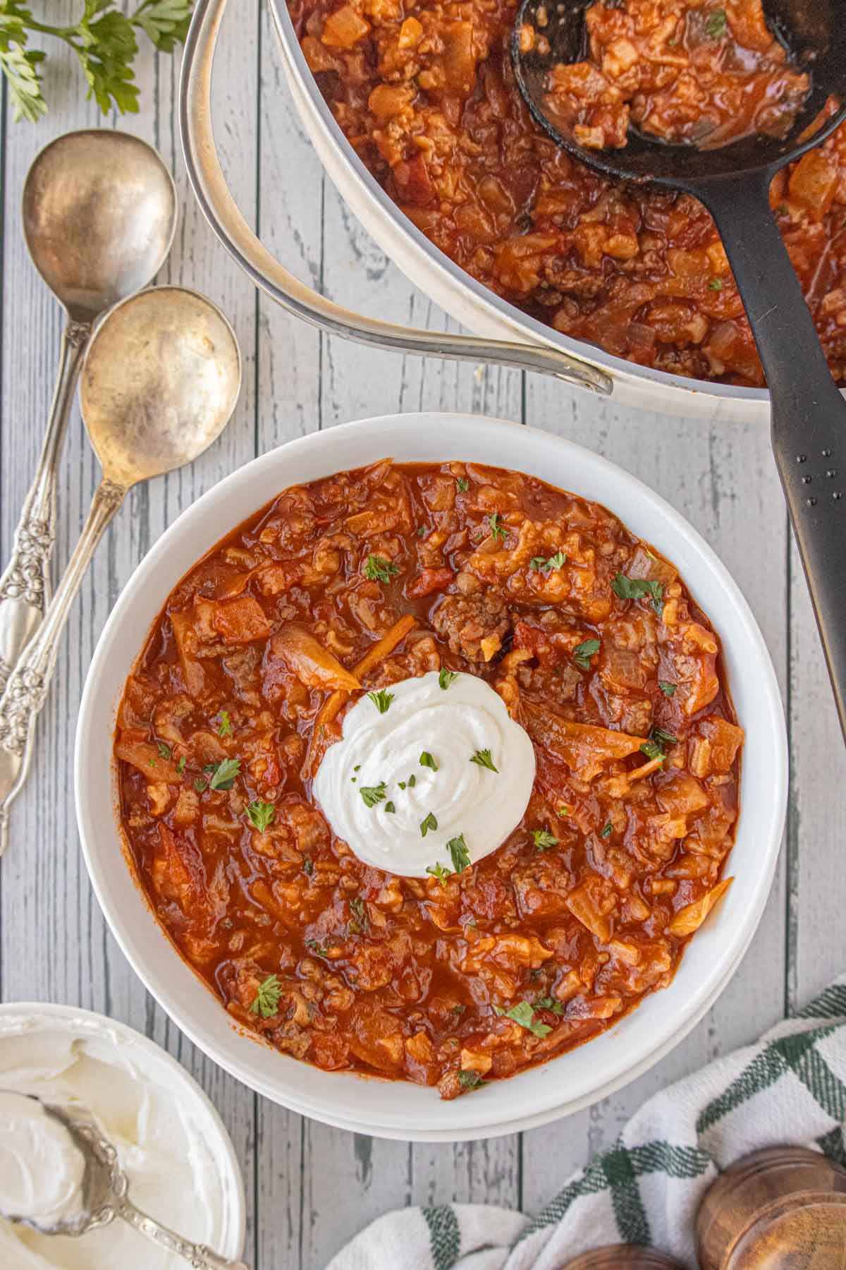 A white bowl filled with cabbage roll soup topped with a dollop of sour cream and fresh herbs, with the pot of soup and vintage silver spoons in the background.