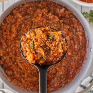 A ladle lifting a scoop of thick, hearty cabbage roll soup from a large pot, showing tender cabbage, ground beef, tomatoes, and rice in a rich red broth.