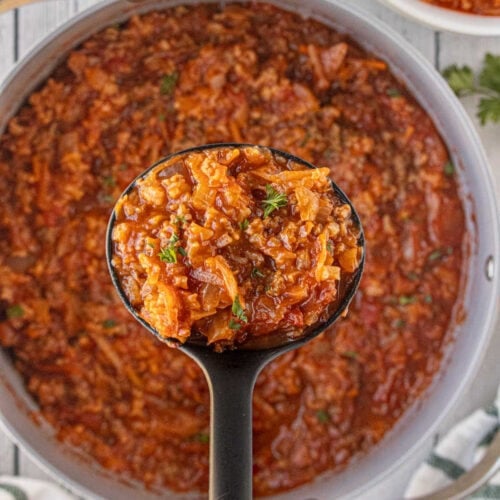 A ladle lifting a scoop of thick, hearty cabbage roll soup from a large pot, showing tender cabbage, ground beef, tomatoes, and rice in a rich red broth.
