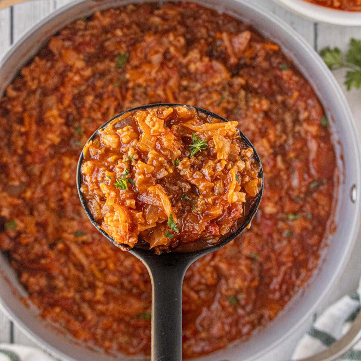 A ladle lifting a scoop of thick, hearty cabbage roll soup from a large pot, showing tender cabbage, ground beef, tomatoes, and rice in a rich red broth.