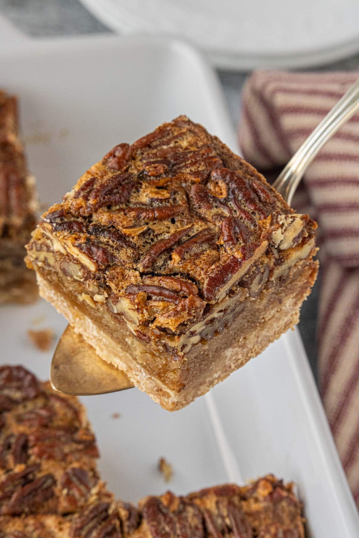 Close-up of a brown butter pecan pie bar lifted from the pan on a vintage pie server, showing the thick gooey pecan filling and buttery shortbread crust.