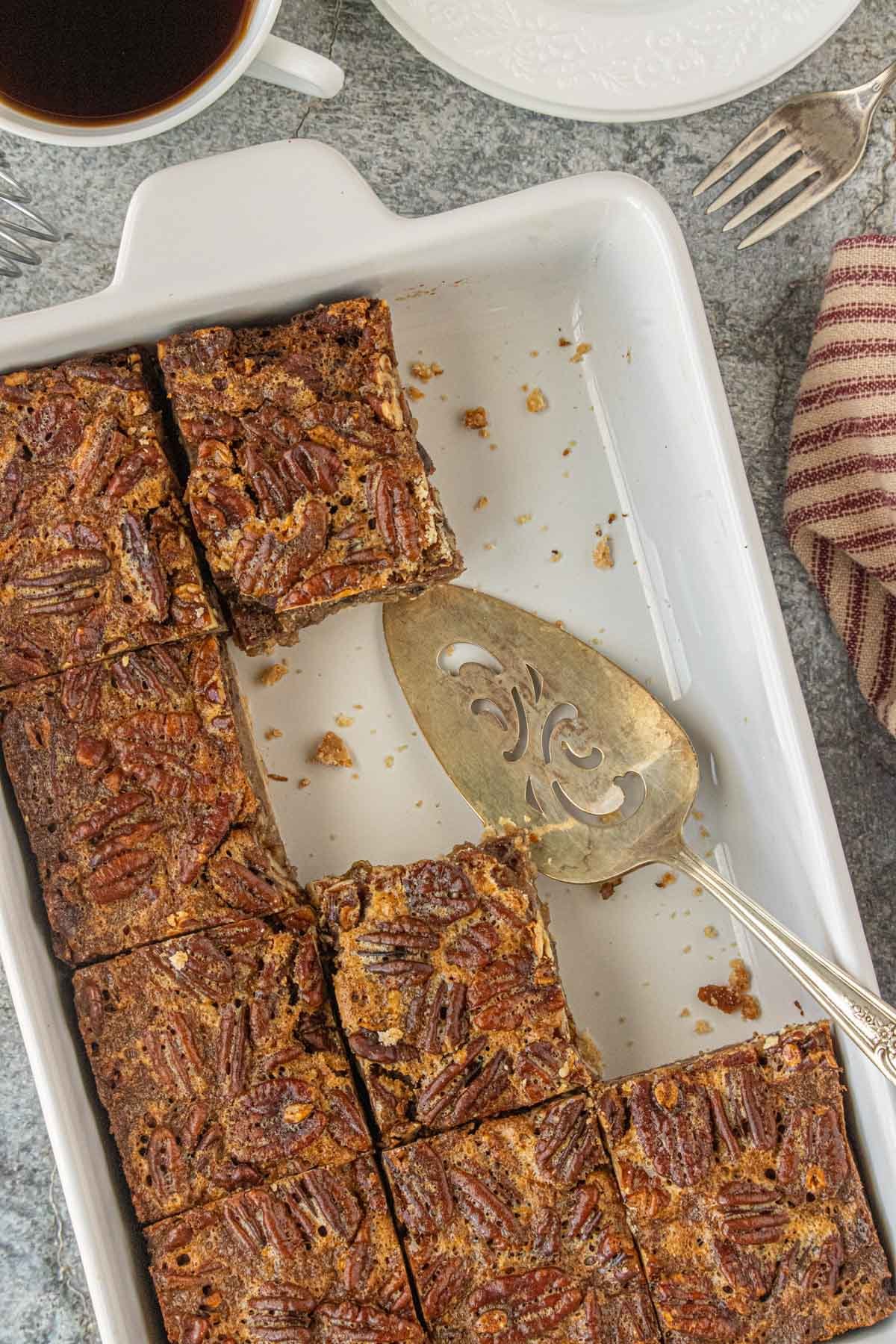 Overhead view of brown butter pecan pie bars in a white baking dish, several squares cut and one missing, with a vintage pie server and a cup of black coffee nearby.