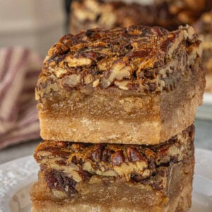 Close-up of two brown butter pecan pie bars stacked on a plate, showing the glossy pecan topping, thick gooey filling, and buttery shortbread crust with visible texture.