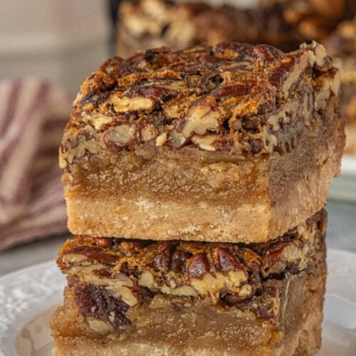 Close-up of two brown butter pecan pie bars stacked on a plate, showing the glossy pecan topping, thick gooey filling, and buttery shortbread crust with visible texture.