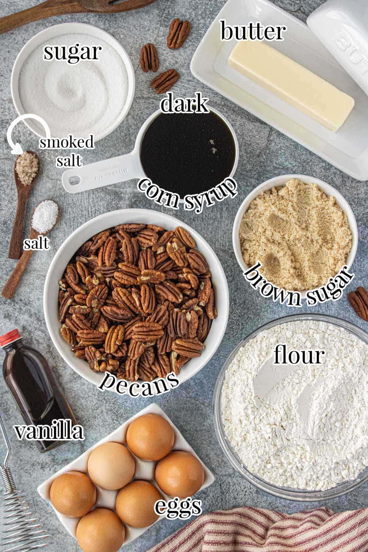 Overhead view of all ingredients for brown butter pecan pie bars arranged on a gray countertop, including pecans, flour, sugar, brown sugar, butter, dark corn syrup, eggs, vanilla, smoked salt, and regular salt, each labeled in text.