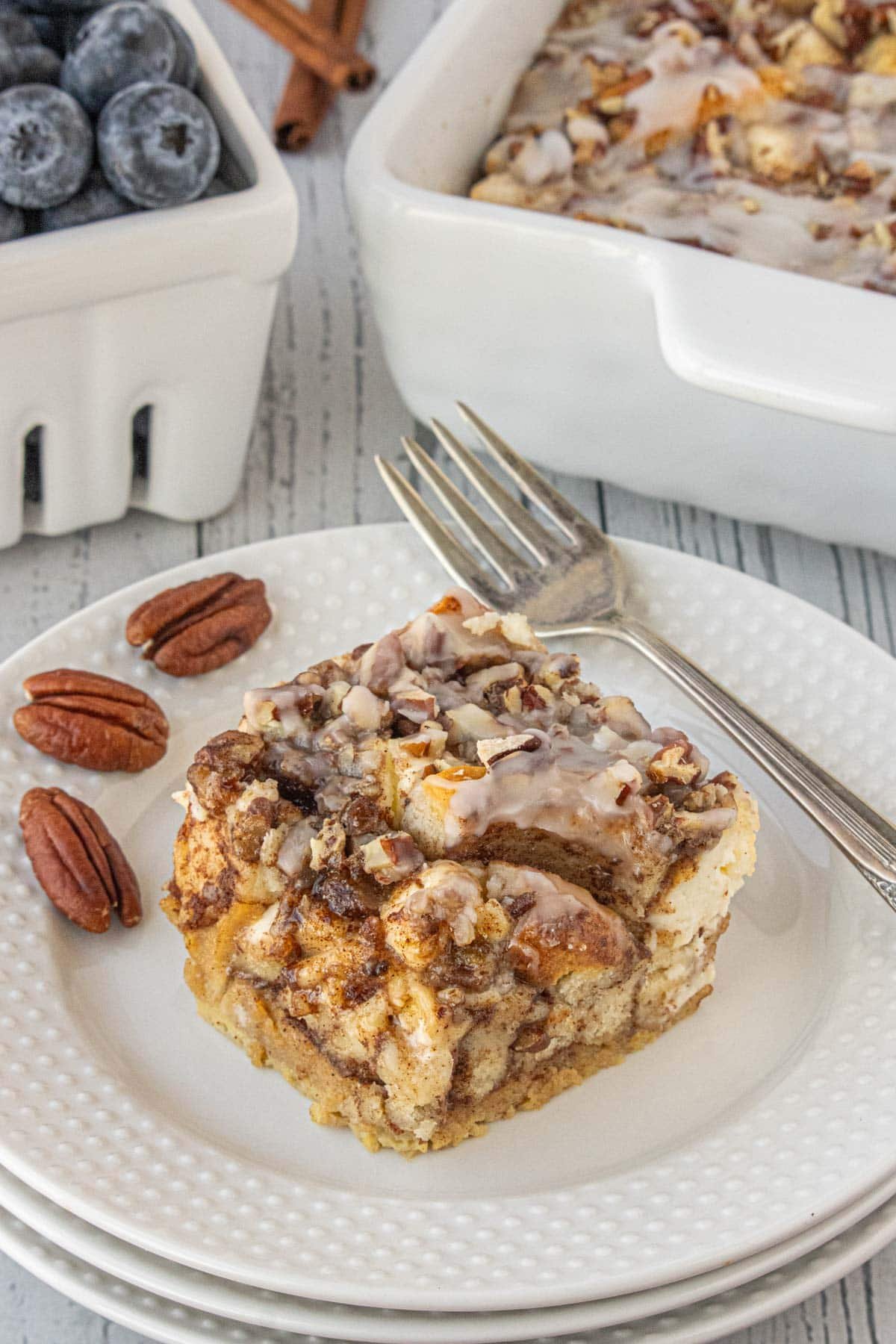 Slice of cinnamon roll breakfast casserole on a white plate with pecans and blueberries, showing swirls of cinnamon, icing drizzle, and soft baked texture.