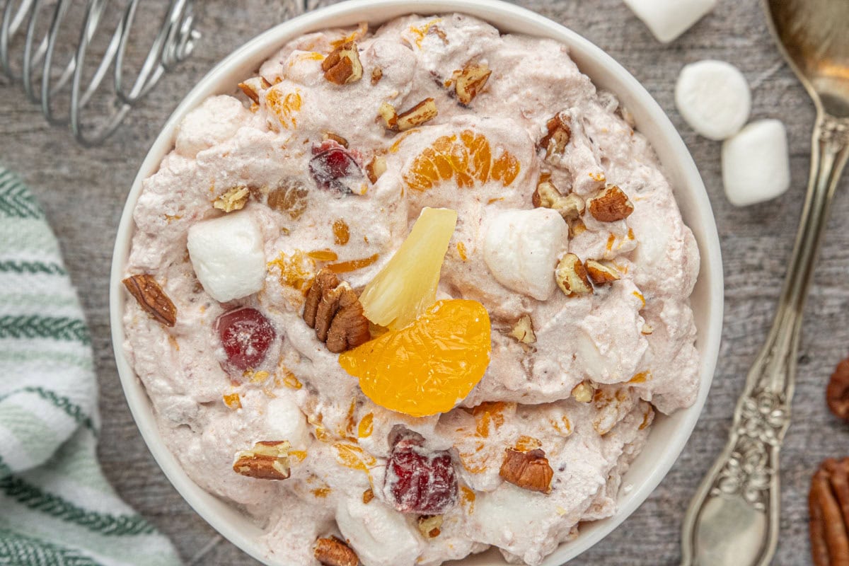 Overhead view of a bowl of cranberry ambrosia salad with mandarin oranges, pineapple tidbits, cherries, pecans, and mini marshmallows folded into a creamy pink mixture.