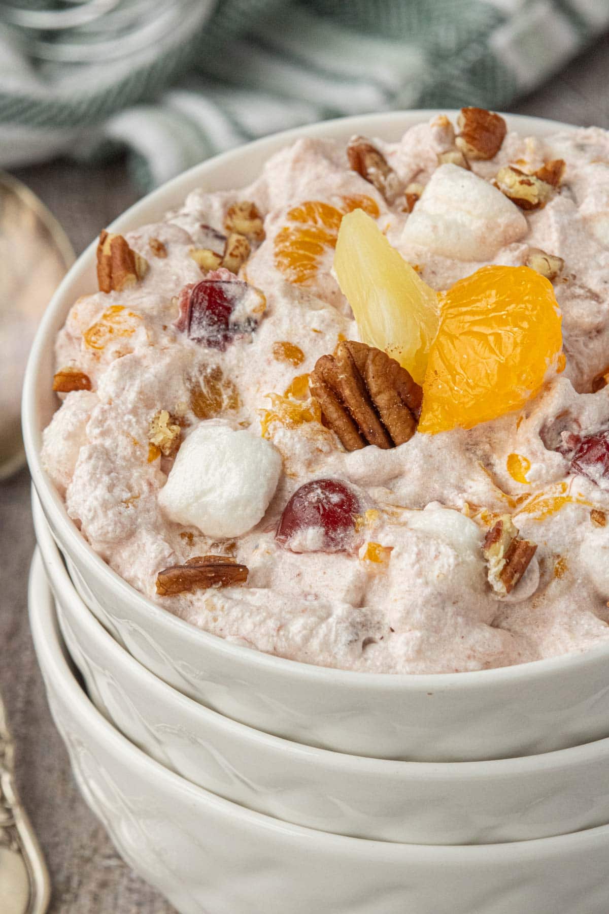 Close-up shot of cranberry ambrosia salad piled in stacked white bowls, showing the creamy pink mixture with mandarin oranges, pineapple, pecans, cherries, and mini marshmallows.