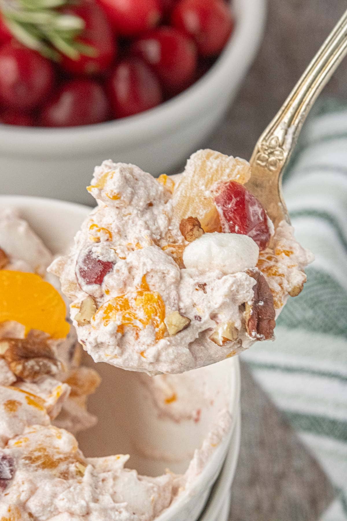 A spoonful of cranberry ambrosia salad lifted from the bowl, highlighting the creamy texture with pineapple, mandarin oranges, cherries, pecans, and mini marshmallows.