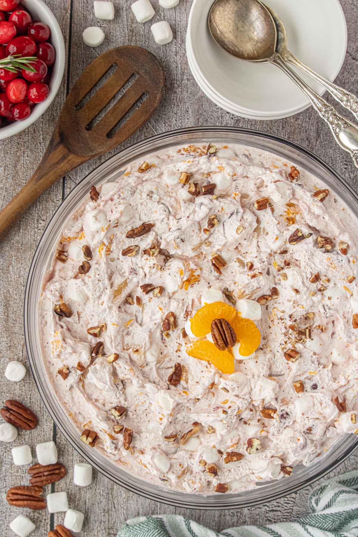 Top-down view of a bowl of cranberry ambrosia salad filled with whipped cream, mandarin oranges, pineapple, pecans, marshmallows, and fresh cranberries, with a vintage spoon and scattered pecans on the table.