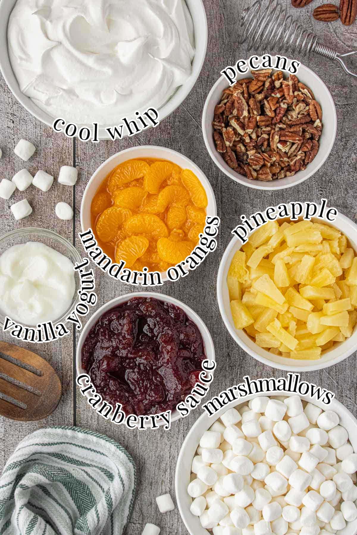 Overhead view of cranberry ambrosia salad ingredients arranged in bowls, including Cool Whip, pecans, mandarin oranges, pineapple, mini marshmallows, cranberry sauce, and vanilla yogurt.