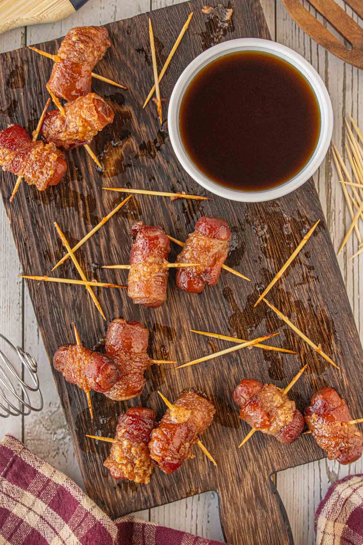 Maple chipotle bacon wrapped little smokies arranged on a wooden board with toothpicks and a small bowl of maple glaze for dipping.
