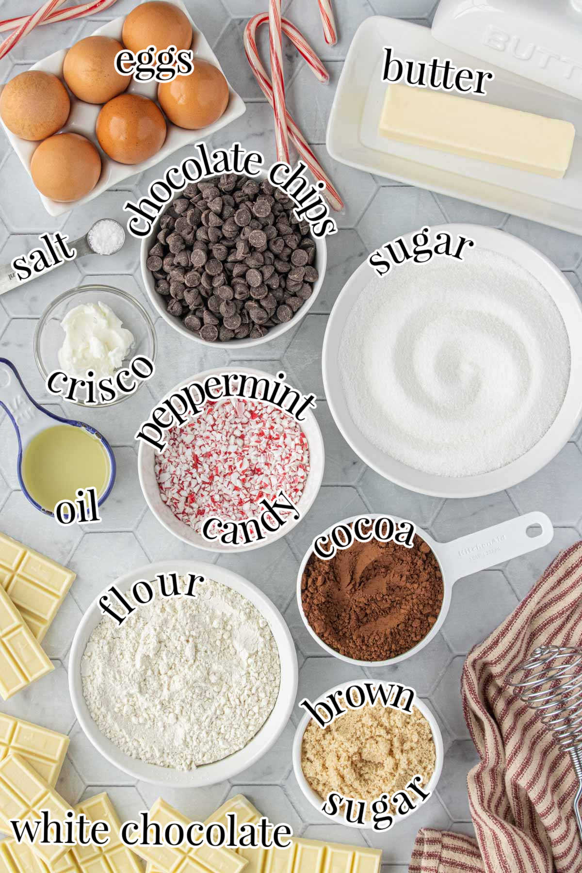 Overhead view of ingredients for peppermint bark brownies, including eggs, butter, sugar, brown sugar, cocoa powder, flour, chocolate chips, white chocolate, crushed peppermint candy, oil, Crisco, and salt, each labeled in the image